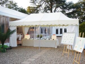 A white marquee tent with a table underneath, set for an outdoor event. The table is adorned with decorative floral arrangements, and two illuminated squares. A large easel holds a seating chart. Trees and a building are visible in the background.