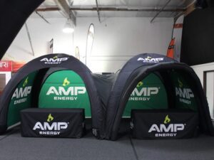 Two black and green AMP Energy branded inflatable tents are set up indoors. They have arched entrances and are placed side by side on a gray carpeted floor. Each tent has a small rectangular display table in front.