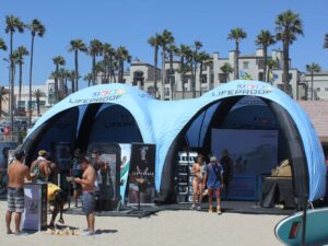 A beach scene with people gathered around a large, blue Lifeproof tent featuring branded displays. Palm trees and buildings are in the background, under a clear blue sky.