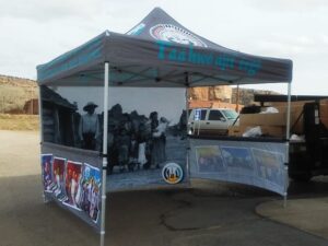 A canopy tent displaying historical black-and-white images of Indigenous people and colorful artwork panels. The tent includes text in a language of Native Americans and is set up in an outdoor setting next to a truck.