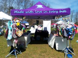A market stall under a purple canopy with the text Made with love in every bite. The stall displays colorful clothes and accessories on racks. Trees are visible in the background.
