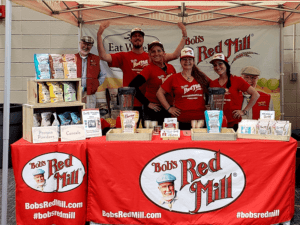 A group of six people in red shirts and caps stand behind a booth featuring Bobs Red Mill products like protein powders and cereals. The booth has a red tablecloth with the Bobs Red Mill logo. Two blenders are also displayed.