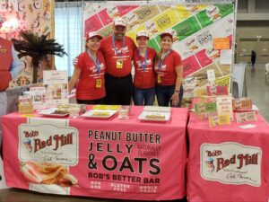 Four people wearing matching red shirts stand behind a display table with Bobs Red Mill products. The table has a pink cloth featuring a peanut butter jelly and oats bar. The backdrop displays a colorful brand image.