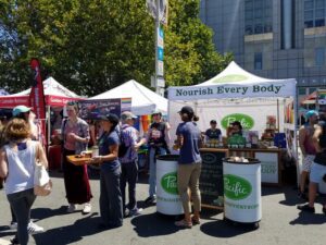 People gather at a colorful outdoor market with various food stalls. A booth with a Nourish Every Body banner is in focus. Attendees are interacting, enjoying the sunny day under leafy trees and clear blue skies.