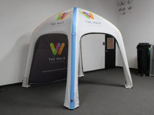A white pop-up canopy tent with The Walk Tuscaloosa logo and multicolored insignia on the side. It stands indoors on a grey carpeted floor, with a black mesh backing partially visible.