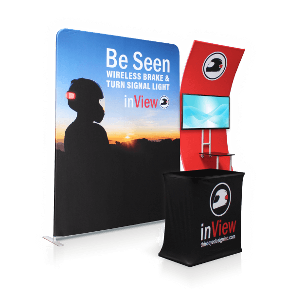 A stylish display booth featuring a large backdrop with a silhouette of a motorcyclist against a sunset and the text "Be Seen Wireless Brake & Turn Signal Light - inView." The setup includes a branded table and TV, creating an engaging booth experience.
