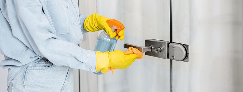 Wearing yellow gloves, a person meticulously cleans a glass door handle with a spray bottle and cloth, ensuring the entrance to the screening tents remains spotless.