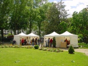 Three 10'x15' V5 Commercial Grade Steel Tents in a park surrounded by trees. People are gathered outside the tents, and a person is serving at a table. The grass is green, and flower beds are in the foreground under a partly cloudy sky.