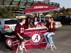 Five people wearing Alabama Crimson Tide jerseys are gathered around a tailgate setup with a branded tent and table. Theyre smiling and talking, with a stadium and parked cars in the background.