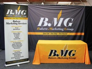 A marketing display featuring a banner and table cover for Bakers Marketing Group. The banner highlights services like advertising and special events. Both the banner and table cover have the company logo and contact details.