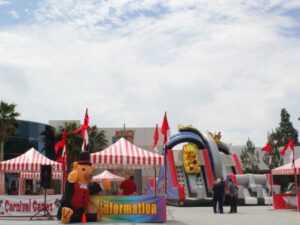A carnival setup with red and white striped tents, an inflatable slide, and a person in a bear costume. Red flags decorate the area, and several people are gathered near the tents. The sky is partly cloudy.