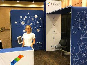 A woman stands at a booth for TRYM, featuring blue displays with geometric designs and icons. The booth showcases a tablet graphic and buzzwords like smart insights and energy control. No visitors are visible in the exhibition area.