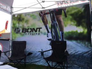 A booth with a Bont Cycling banner displaying a cyclist on a road bike. Two empty black folding chairs are in front of the banner, and a water bottle is on the ground between them. The scene is under a canopy tent.