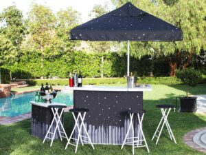 Outdoor bar setup with a starry night-themed umbrella and counter, surrounded by folding stools. Wine bottles and glasses are on display. The scene includes a grassy area and a swimming pool in the background on a sunny day.