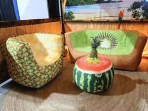 A tropical-themed room with a pineapple-patterned chair, a kiwi-printed sofa, and a watermelon-shaped ottoman. A faux pineapple decor is placed on the ottoman, and a vibrant parrot mural is in the background.