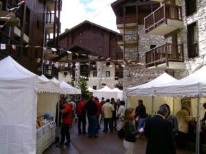 An outdoor farmers market in an alpine village features white tents and wooden buildings. People gather, browsing stalls with goods on display. Red and white bunting is strung overhead, creating a festive atmosphere.