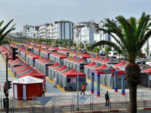 A vibrant farmers market scene unfolds with rows of stalls covered by red and gray canopies. Palm trees line the street, and people walk among the booths. White buildings with balconies stand tall in the background under a clear sky.
