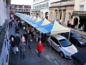A bustling street market with people strolling past farmers market tents covered by yellow and blue striped canopies. A white HELPING HANDS car is parked near the stalls. Buildings line the street, and a street performer is visible among the crowd.