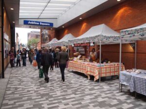 A bustling farmers market in Jubilee Square buzzes with people browsing stalls. These stalls, beneath white tents, display a variety of colorful products. The alley is paved with patterned tiles, and a Mister Tuckshop sign is visible amidst the lively scene.