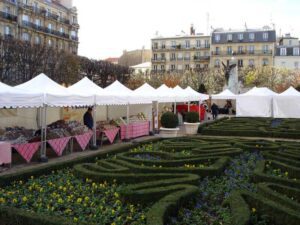 A series of white farmers market tents line a walkway, with tables displaying goods covered in red checkered cloths. In the foreground, manicured hedges and flowers create an ornate garden pattern, set against the backdrop of buildings.