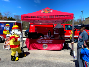 A fire safety event features a red tent with Union Fire District Fire Prevention written on it. A person in a Dalmatian fire mascot costume stands nearby. A red fire truck is in the background, and several people are gathered around the tent.