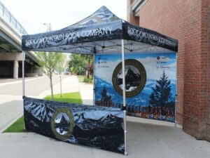 A blue and black tent promoting Rocky Mountain Soda Company is set up on a sidewalk. The tent features a mountain and pine tree design, with labels indicating vegan, gluten-free, non-GMO, and caffeine-free options.