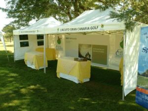 Two white school tents are set up outdoors for the Circuito Gran Canaria Golf event. Each tent features tables covered with yellow cloths. Banners display event branding, while nearby trees offer shade on the grassy area.