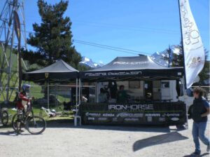 A cyclist stands next to a mountain bike in front of an Iron Horse booth, complete with banners and high peak pop-up tents. The backdrop features a mountainous landscape with snow-capped peaks and a clear blue sky, as people gather around the lively booth.