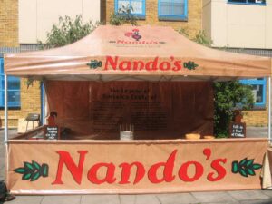 A Nandos outdoor stall, reminiscent of modern food concession tents, features a beige canopy adorned with the iconic Nandos logo. Set against a charming brick building, the counter brims with enticing promotional materials and an array of flavorful condiments.