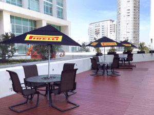 Outdoor patio with round tables and chairs under black umbrellas featuring the Pirelli logo. The area has wooden decking and is surrounded by modern buildings, plants, and sculptures under a clear sky.