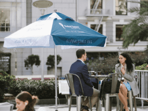 Two people seated at an outdoor table under a blue and white umbrella with Global Genes and #RAREintheSquare printed on it. They are engaged in conversation, with trees and a building facade in the background.