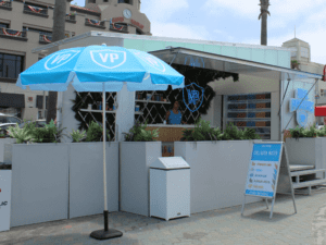 A modern outdoor kiosk with a light blue VP-branded umbrella. The setup includes a counter, decorative plants, and a menu board listing drinks. A person is visible behind the counter. Buildings and palm trees are in the background under a cloudy sky.