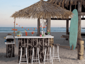 Tiki bar on a sandy beach under a straw umbrella, surrounded by barstools with a bamboo pattern. Colorful drinks sit on the counter. A surfboard and a string of lights add to the tropical vibe, with the ocean and sky in the background.