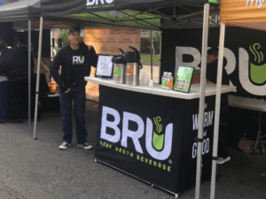 A man stands next to a booth with a banner displaying BRU Bone Broth Beverage. The booth has a counter with thermoses, cups, and promotional materials. There are other vendors nearby under canopy tents.