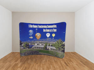 A curved display banner with the text 5 Star Homes Transforming Communities, One Home at a Time. The graphic features a landscape with hills and trees and hot air balloons in the sky. The banner is set against a plain room with wooden floors.