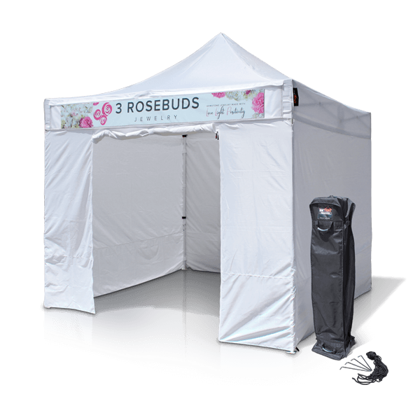 A white pop-up tent with the text 3 Rosebuds Jewelry on the front valance. It features decorative floral designs. A carrying case for the tent lies next to it on the ground.