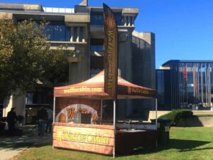 A food concession tent with a canopy labeled Waffle Cabin is set up outdoors on a sunny day. The tent boasts orange and brown hues, featuring a waffle pattern. A tall flag displays the same name, while buildings and greenery serve as the perfect backdrop.