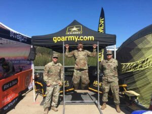 Three people in military uniforms stand under a goarmy.com tent. One person is doing pull-ups on a bar, while the other two stand nearby smiling. The background includes another tent and clear blue sky.