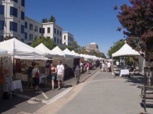 Outdoor street market with vendors in white tents lined along a road. People walk between the stalls under a clear blue sky. Buildings and trees are visible in the background.