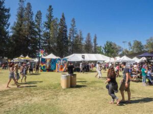 People attend an outdoor festival on a sunny day, with white tents and vibrant artwork displayed. Trees line the background, and attendees walk across the grassy area, enjoying the lively atmosphere.