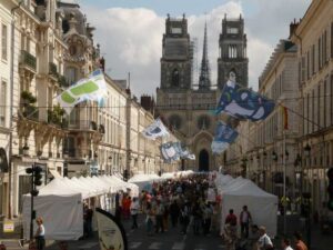 A bustling street fair with white tents lines a wide road, leading to a large, historic cathedral with twin towers in the background. Colorful flags hang above the crowd, creating a festive atmosphere under a partly cloudy sky.