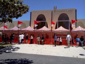 A line of people stands outside a convention center with red and white striped tents labeled Carnival Games. Red flags adorn the tents, and the sun casts shadows on the pavement.