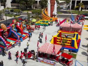 A vibrant outdoor carnival with colorful inflatable games, such as slides and obstacle courses, surrounded by people. Red and white striped tents house more activities and vendors. The area is lively and festive.