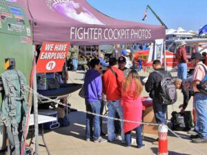 People gather under a tent labeled Fighter Cockpit Photos at an outdoor event. A sign offering earplugs for $1 is visible. Nearby, there are aircraft and a table with informational materials. The sky is clear and blue.