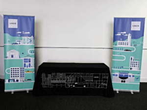 A table covered with a black cloth featuring a white outline of a cityscape is flanked by two vertical banners. The banners have a similar cityscape design with roads, cars, and buildings, and both display the Uber logo at the top.