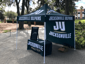 A promotional tent for Jacksonville Dolphins athletics is set up outdoors. The tent features the JU logo and Jacksonville Dolphins branding on its canopy. A table under the tent is covered with a cloth that also displays Jacksonville Athletics.