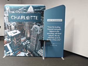 A display featuring the Charlotte skyline with the word CHARLOTTE and crown logo. Beside it, a panel reads: Meet in Charlotte and a quote about the city’s vibrant metropolitan life by Southern Living. A small black table is in front.