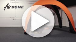 A large play button overlays a video still showing an orange and black custom inflatable tent inside a spacious room with grey carpeting. The word AirDOME is displayed on the wall in stylized lettering.