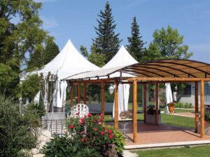 A garden with a wooden pergola on a brick patio, surrounded by greenery and flowers. Two white high peak pop-up tents stand nearby under a clear blue sky.