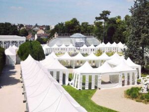 A series of high peak pop-up tents adorns the outdoor garden, nestled among trees and shrubs. The landscaped paths and open spaces create a tranquil setting with a city skyline backdrop under the clear blue sky.